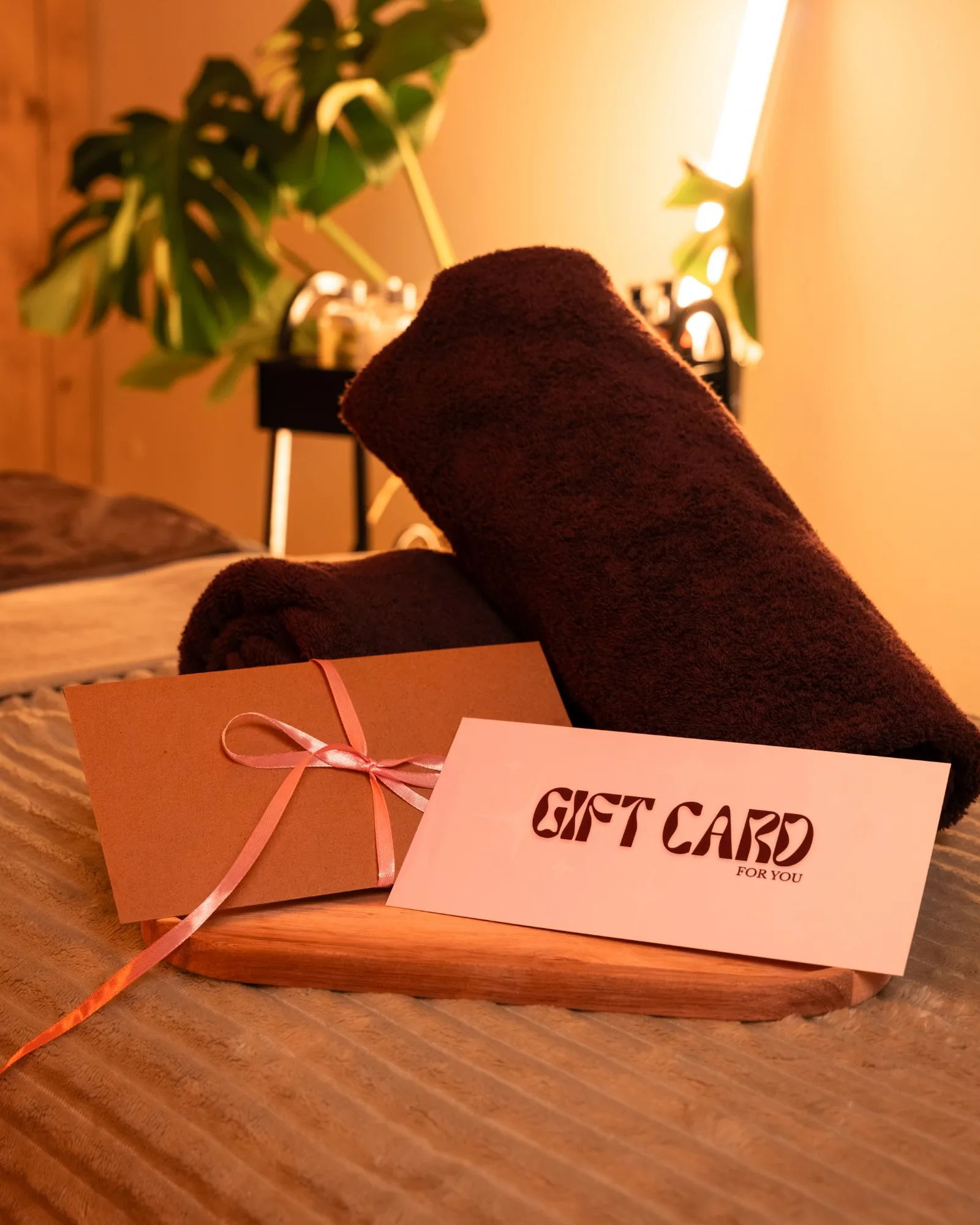 Gift card na masaż w kopercie z różową wstążką, obok zwinięty brązowy ręcznik